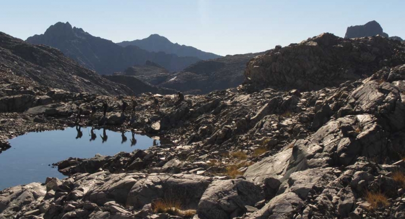 A group of students hike alongside an alpine lake, which is reflecting their presence. The lake is surrounded by rocks, and there are mountains in the background. 